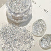 Clear acrylic rhinestone flakes in a small clear jar, with loose silver-clear stones scattered on a white surface and two polished stone-like pieces beside it. Chinese text appears in the top left and bottom right, and part of a printed design is visible in the background.