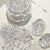 Clear acrylic rhinestone flakes in a small clear jar, with loose silver-clear stones scattered on a white surface and two polished stone-like pieces beside it. Chinese text appears in the top left and bottom right, and part of a printed design is visible in the background.