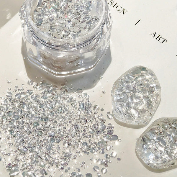 Clear acrylic rhinestone flakes in a small clear jar, with loose silver-clear stones scattered on a white surface and two polished stone-like pieces beside it. Chinese text appears in the top left and bottom right, and part of a printed design is visible in the background.